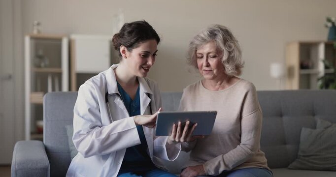 Female doctor sits with senior 70s woman patient, showing health information on tablet, explains test result or new digital telemedicine application use during home visit. Consultation, medical care
