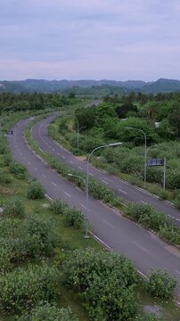 Aerial Winding Road Through Scrub And Bushes, Empty Lanes Curve Toward Distant Hills, Lampposts And Signage Align Roadside, Overgrown Verges And Quiet Cloudy Sky, Conservation Mapping Tone