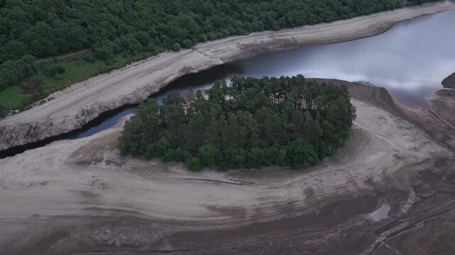 A lush forest thrives on a parched island, surrounded by dry, cracked earth bordering a vast body of water.