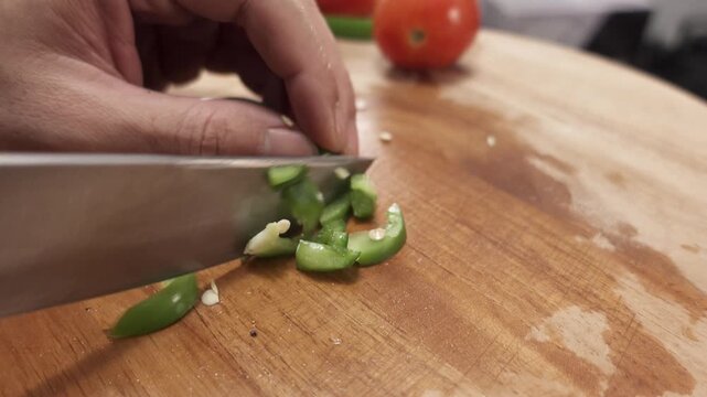 Close-up of a hand chopping green bell pepper into small pieces with a kitchen knife on a cutting board, preparing fresh vegetables for cooking