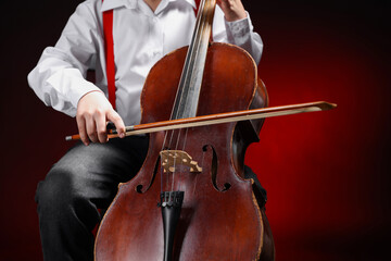 Little boy playing violoncello on dark red background, closeup