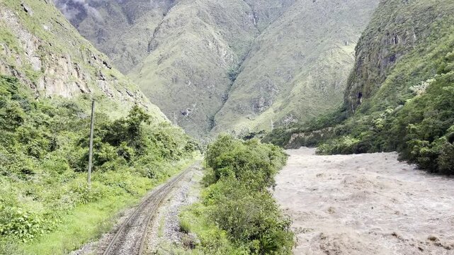 Rear view of a tourist train slowly climbing the Andean railway toward Machu Picchu beside a swollen muddy river in flood. The soundtrack captures the rhythmic rail clack of the tracks. Filmed in Peru