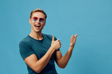 Cheerful young man wearing sunglasses and a blue T-shirt showing thumbs up gesture against a plain...