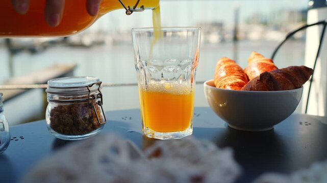 Hand pours fresh orange juice into a glass on an outdoor table with croissants and granola, overlooking a sunny marina. Captures a refreshing breakfast moment by the water
