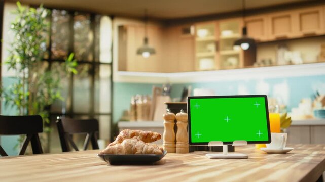 Chroma key display on tablet in bright spacious kitchen with breakfast table, wooden furniture with kitchenware and decorative items highlight a modern apartment design with no people.