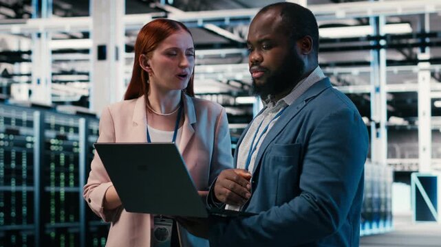 Optimizing electronics, laptop and manager teaching intern in server farm during induction. Monitoring system performance, notebook and team leader with trainee in data center, camera B