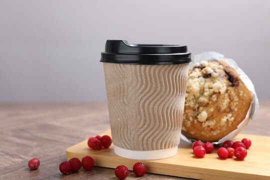 Tasty muffin, coffee in takeaway cup and cranberries on wooden table against grey background, closeup