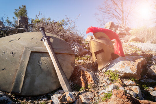 Ancient Greek Spartan helmet with red plume, sword and shield resting against stone ruins outdoors