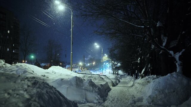 Snowy night street with huge snowdrifts and bare trees under bright city lights