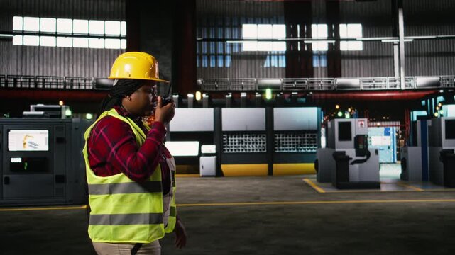Woman engineer, portable walkie-talkie, speak with colleagues, walking across warehouse, worker supervising machinery hall workstations. Wireless radio transceiver near sparks from welding.