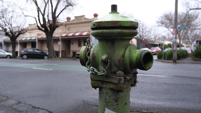 A vintage and weathered green pillar fire hydrant stands on a residential street corner in Fitzroy, against a backdrop of historic Victorian-era terrace houses in suburban Melbourne Australia. 