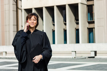 30s Asian woman in black academic gown standing in plaza holding mortarboard using pink smartphone © wavebreak3