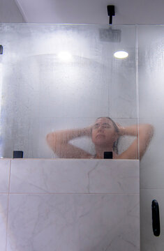 Mujer hispana mexicana tomando una ducha caliente como parte de la higiene personal ba&ntilde;o diario de la cultura de Mexico. ba&ntilde;o con luz blanca y puertas de cristal, ducha caliente, vertical