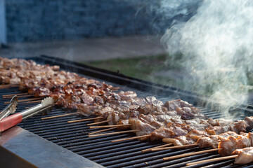 A barbecue utensil with red handles lies on a grill with a row of boneless, skinless chicken cooking. The Korean chicken and spices are on wooden skewers, grilling with smoke rising from the heat.