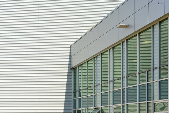The corner of a large grey building with metal composite panels and a row of commercial glass windows. There's a larger white exterior metal-clad siding wall of a building in the background.