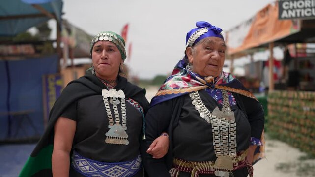 Two smiling Mapuche women in traditional silver jewelry and clothing are walking and talking together