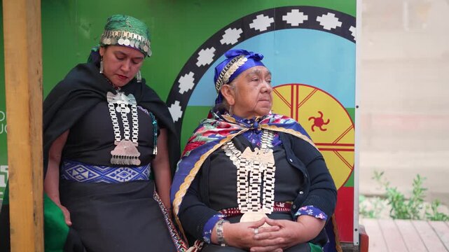 Two Mapuche women in traditional dress sitting in front of the indigenous flag of Chile