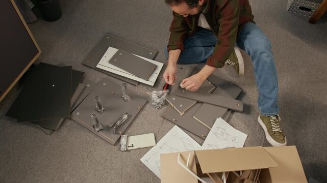 Full top view of mature man in casual jeans and shirt assembling flatpack chipboard locker on floor at home, inserting fixtures and using screwdriver tool, box and instructions nearby