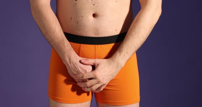 Urology. Man in orange underwear showing thumbs down and covering his groin with hands on purple background, closeup. Camera moving down