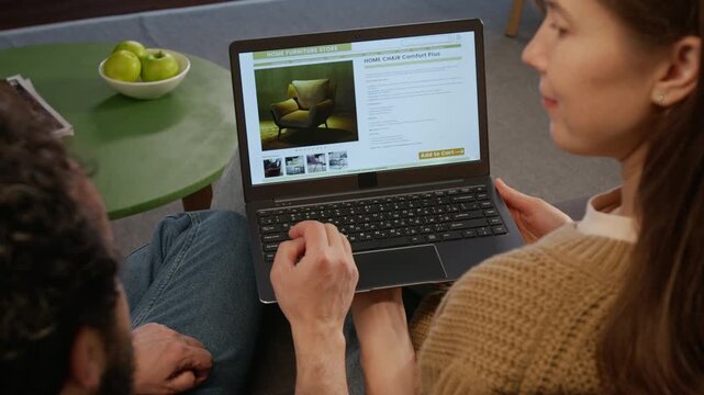Over-shoulder shot of couple sitting together on sofa, looking at photographs of armchair in online furniture store on laptop screen, discussing design and style for home renovations