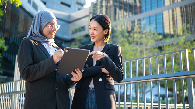 Portrait of two smiling diverse Asian businesswoman, one wearing hijab, discussing work on a tablet together outdoors against a modern office building and green trees background.