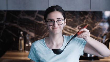 Young woman holding ladle in kitchen smiling at camera, A girl holds a ladle with ready soup and...