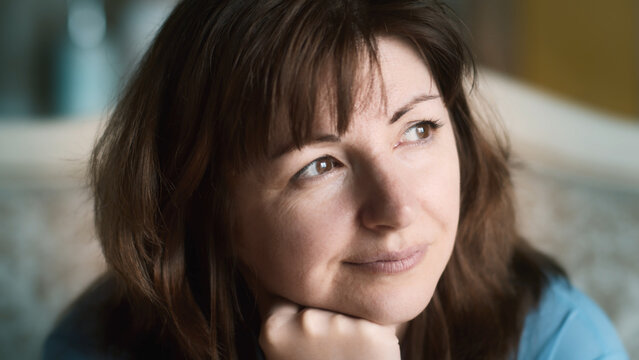 Thoughtful middle-aged woman looking away with hand resting, happy woman, light from the window falling on her face