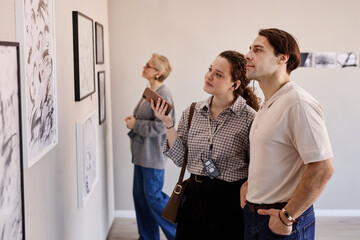Obraz premium Young Caucasian woman and young Caucasian man standing side by side observing framed artwork on gallery wall, middle aged Caucasian woman in background examining other pieces