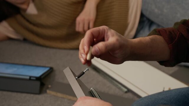 Medium shot of anonymous woman lying on floor carpet, chatting to man assembling flatpack drawer, inserting dowels and tightening bolts with screwdriver, instruction on digital tablet