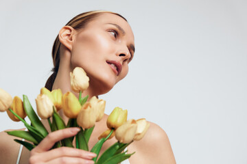 Naklejka premium A young woman with clear skin holds a bouquet of yellow tulips close to her chest while looking thoughtfully to the side against a plain light background, beauty concept.