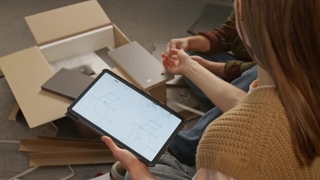 High-angle over-shoulder shot of unrecognizable woman with instruction manual on tablet assisting man assembling knockdown drawer at home, pointing to fixtures and bolt holes on panel