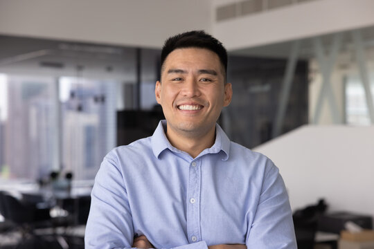 Confident young Asian businessman posing for headshot portrait in corporate office enjoying successful work achievements or creating professional profile picture for social media and career platforms