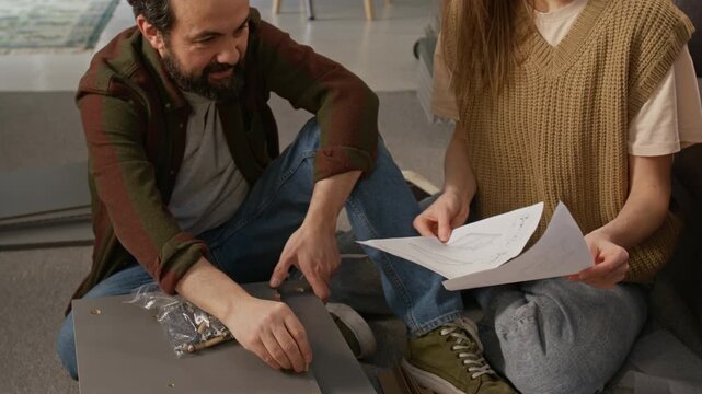 Medium full dolly shot of frustrated mature Caucasian man inserting wooden fixture dowels on cabinet panel while talking to wife holding assembly instructions, while making knockdown furniture at home