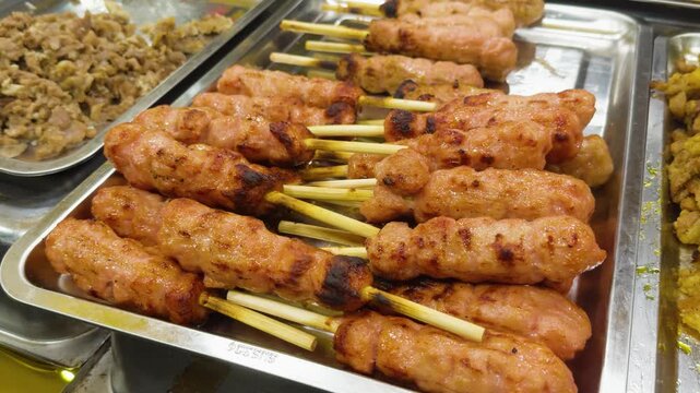 Nem nuong, a popular vietnamese street food, displaying grilled pork skewers prepared with aromatic lemongrass, resting on a metal tray alongside other fried dishes in an authentic market setting