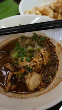 Traditional Thai boat noodles with thick spicy pork broth, meatballs, and liver in ceramic bowl with chopsticks