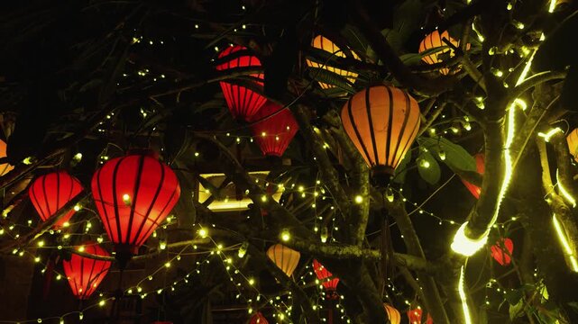 Vietnamese silk lanterns in red and orange hang from tree branches amid warm white string lights, casting a glowing, festive ambiance over hoi ans historic evening streetscape