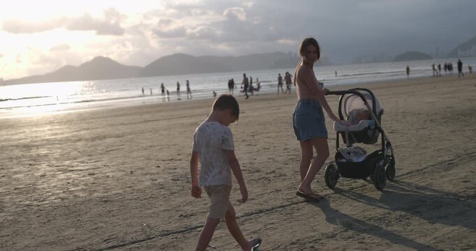 Mother pushing baby in stroller along beach while young son follows behind upset family vaction