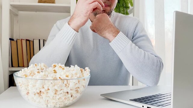 Man in a light blue sweater watches a movie on a laptop at home with popcorn. Cozy atmosphere: books, plants, and lighting. Modern home entertainment blends technology and tranquility