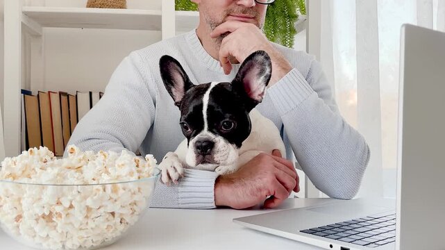 Man in a light blue sweater watches a movie on a laptop at home with his dog, enjoying popcorn. Cozy atmosphere: books, plants, and soft lighting. Modern home entertainment blends technology and tranq
