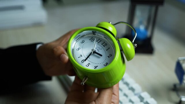 Green twin-bell alarm clock on desk, Hand holding a bright green twin-bell alarm clock on a desk with keyboard and hourglass