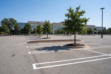 A spacious empty asphalt parking lot sits vacant in front of a modern commercial office building surrounded by green trees