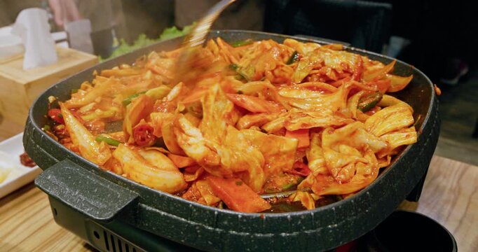 The process of cooking Korean-style spicy stir-fried spring chicken on an iron plate involves stir-frying tender chicken meat, chewy rice cakes, and kimchi in a bright red spicy sauce, releasing an en
