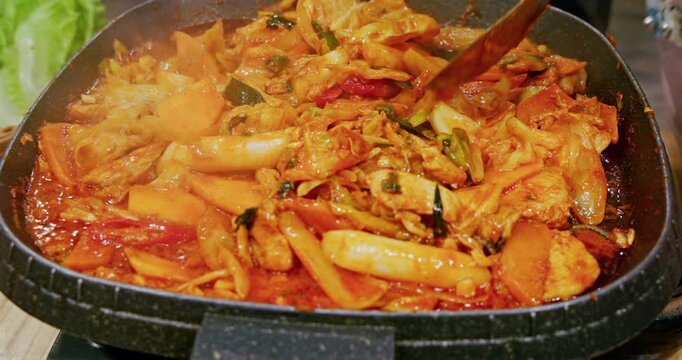 The process of cooking Korean-style spicy stir-fried spring chicken on an iron plate involves stir-frying tender chicken meat, chewy rice cakes, and kimchi in a bright red spicy sauce, releasing an en