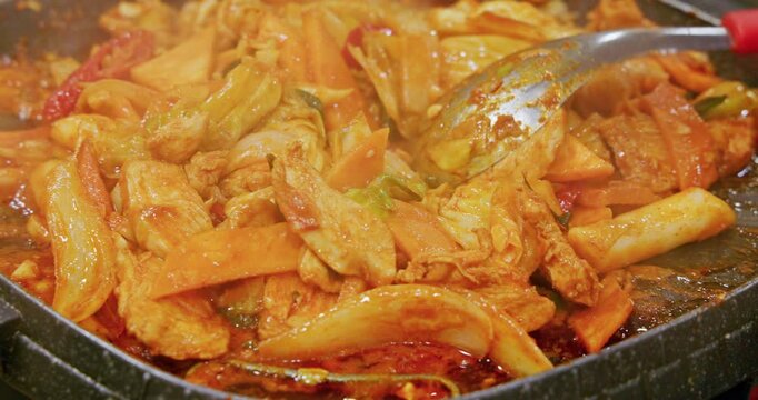 The process of cooking Korean-style spicy stir-fried spring chicken on an iron plate involves stir-frying tender chicken meat, chewy rice cakes, and kimchi in a bright red spicy sauce, releasing an en