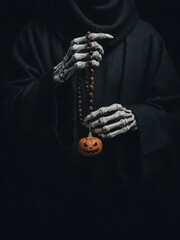 Skeleton hands in black hooded robe holding dark prayer beads with small carved Halloween pumpkin jack-o-lantern.