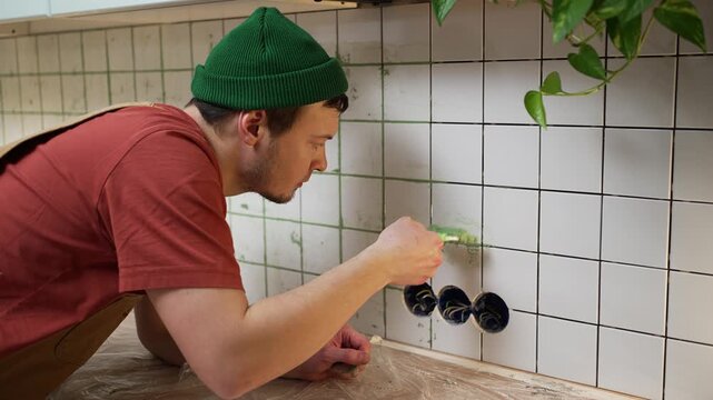 Professional builder meticulously applying colored grout to the seams of a white tiled kitchen backsplash, carefully using a brush to fill the gaps between the ceramic squares around the sockets
