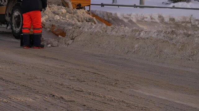 Yellow municipal snow plow actively clearing a city street covered with a mixture of snow and sand, pushing the icy slush to the roadside to improve traffic safety and accessibility