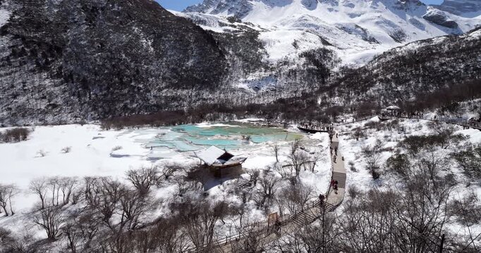 Beautiful panoramic view of snow covered Huanglong National Park in China, with visitors walking along wooden boardwalk beside famous turquoise travertine calcite pools during winter.
