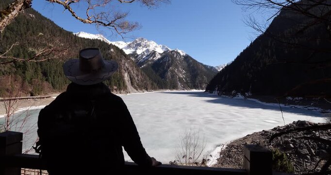 Looking to frozen Long Lake, camera move back and reveal silhouette of tourist man with hat admiring stunning winter view. Dark mountains and snow capped peaks at background