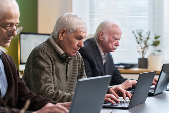 Group of senior Caucasian men sitting in classroom setting using laptop computers, participating in computer literacy course, focusing on learning digital skills together